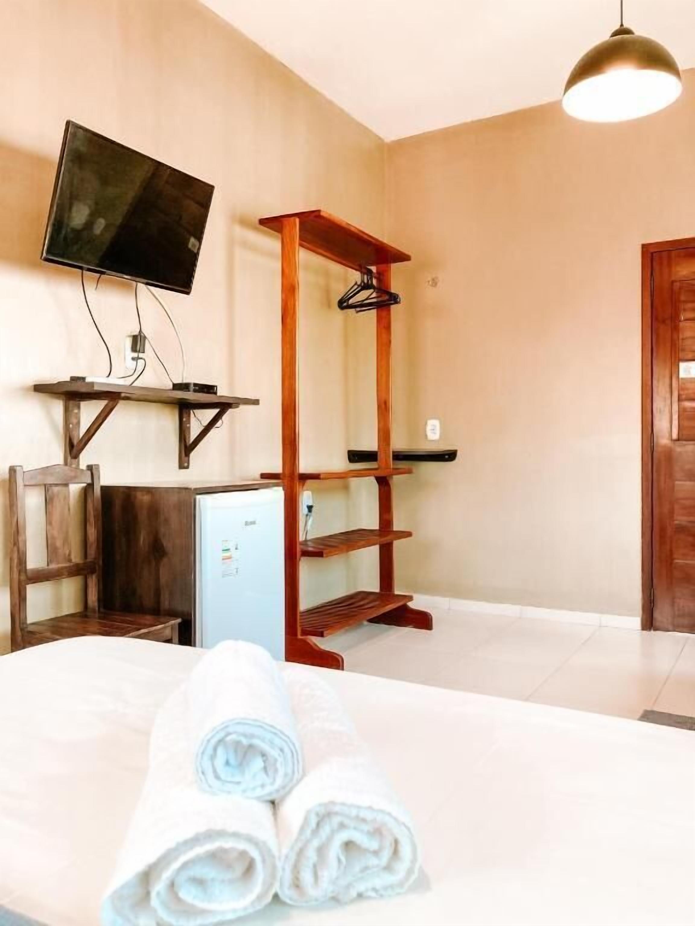 Una camera d'albergo con un letto, una TV a muro, una scaffalatura in legno, un piccolo frigorifero e una porta.