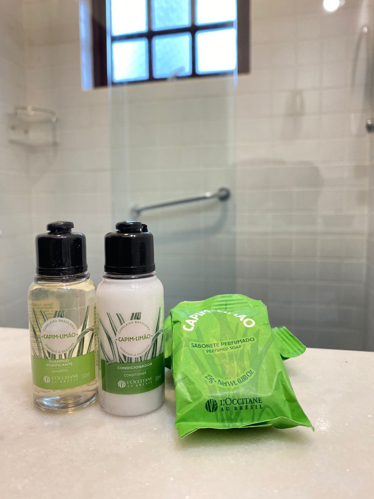 Deux flacons de shampoing et d’après-shampoing Capim-Limão, et un sac vert contenant du savon L’OCCITANE AU BRASIL, posés sur le comptoir d’une salle de bain.