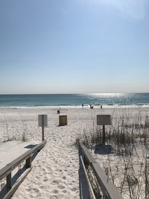 A sandy beach with a wooden walkway leading to the ocean. There are signs indicating no trade ordinance and a warning about the beach.