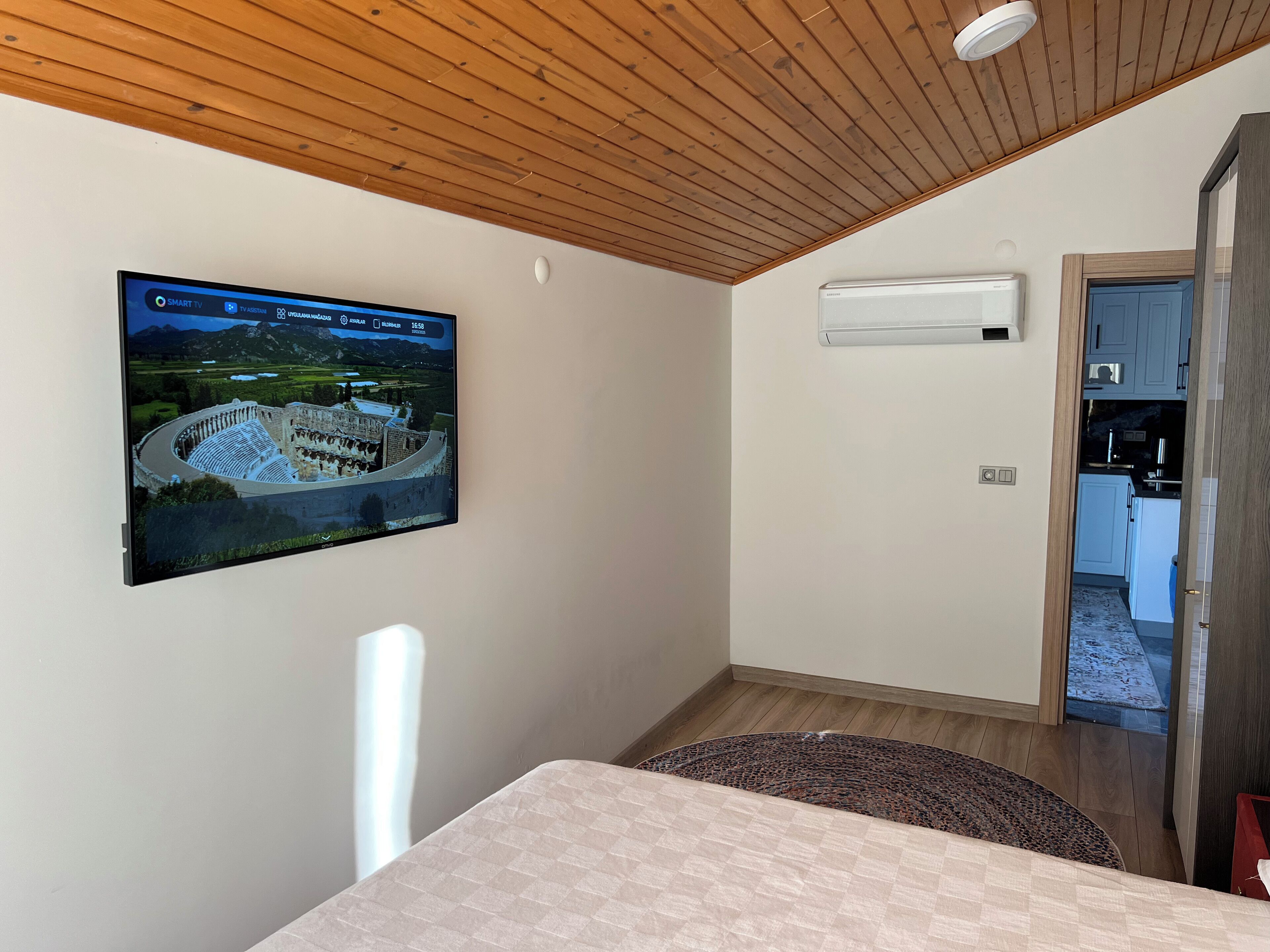 Een kamer met een houten plafond, een tv met daarop een landschapsbeeld, een airconditioning en uitzicht op een keuken.