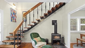Una escalera con peldaños de madera y barandillas negras, un sillón verde con cojín, una mesita y un estufa de leña.