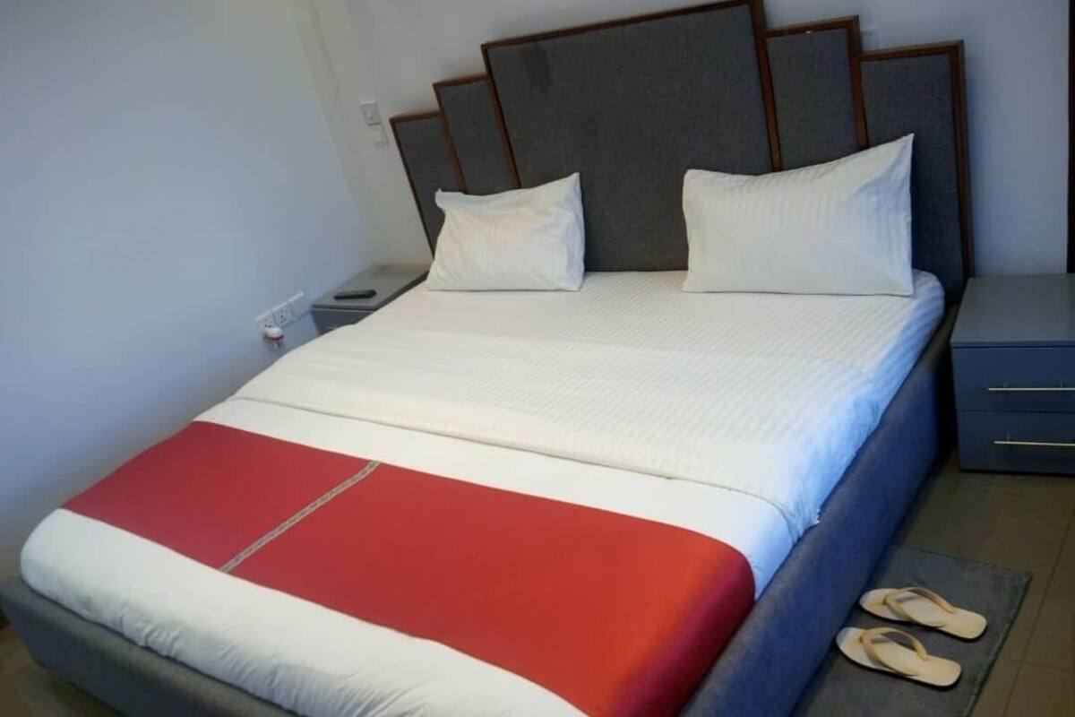 A neatly made bed with a red and white bedspread, two pillows, and a pair of slippers on the floor.