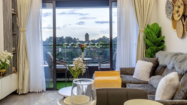 Un salon moderne avec un canapé, une table basse et un coin repas avec des chaises. On aperçoit un balcon avec une porte vitrée, agrémenté d’une table avec des fleurs et de chaises.
