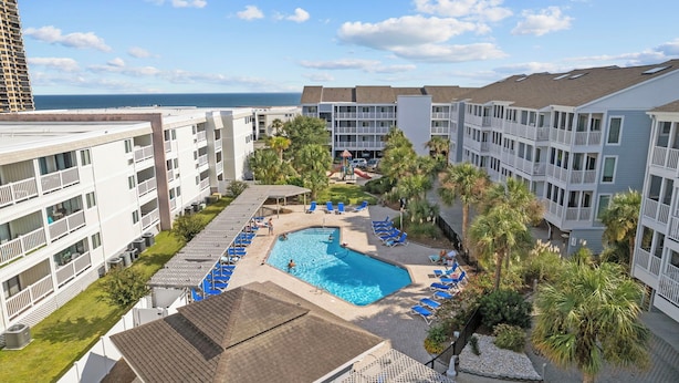 A hotel with a central swimming pool surrounded by lounge chairs, palm trees, and a view of the ocean.