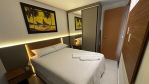 Un lit bien fait, avec du linge de lit et des serviettes blanches, une tête de lit en bois, une table de chevet, un miroir et une armoire en bois.