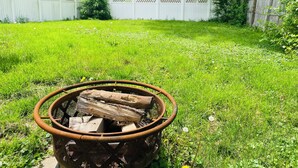 A backyard with a metal fire pit containing logs, surrounded by a white fence and lush greenery. - Spacious 3BR Home w/ 8 Beds, Big Yard & Parking – Near Canton & Hall of Fame (Massillon)