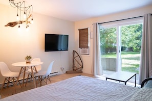 A modern bedroom with a large bed, a flat-screen TV, a dining table with chairs, and a view of a garden through a sliding glass door.