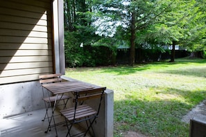A wooden table and chairs set up on a porch with a grassy yard and trees in the background.