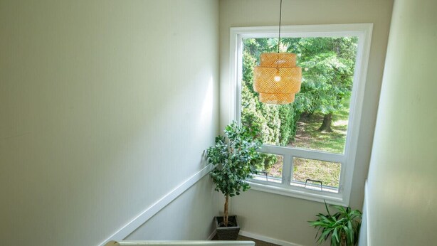 Un escalier avec une rampe blanche, une lampe suspendue en osier et une plante en pot près d’une fenêtre donnant sur un paysage verdoyant.