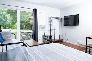 A modern bedroom with a bed, a desk, a chair, a TV, and a balcony with a view of greenery.