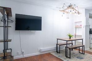 A modern living room with a flat-screen TV mounted on the wall, a wooden dining table with benches, a hanging light fixture, and a potted plant.