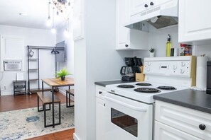 A modern kitchen with white cabinets, a stove, and a coffee maker. There is a wooden table and a plant in the dining area.