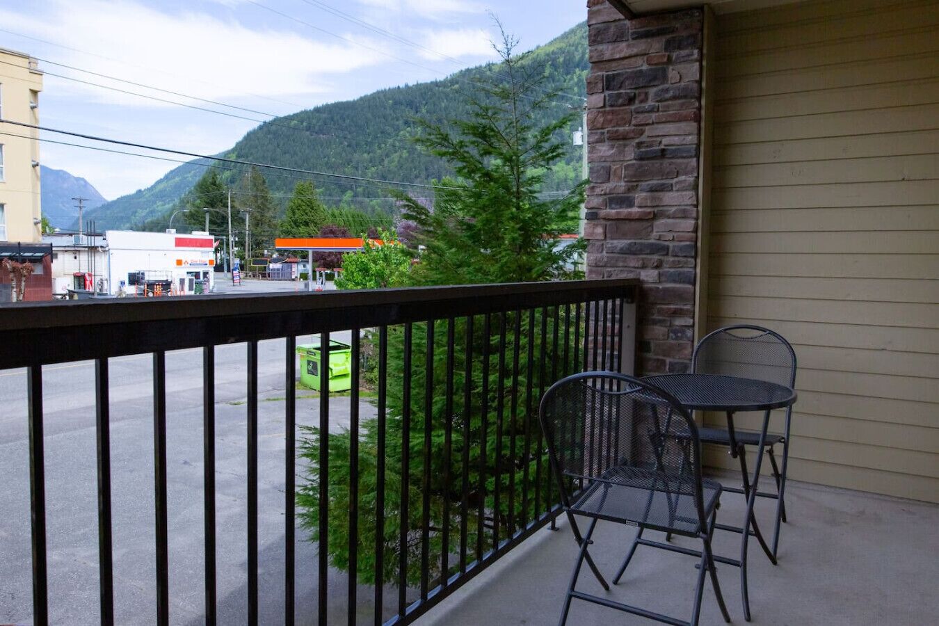 A balcony with a metal railing, two metal chairs, and a view of a street with buildings and a mountain in the background.
