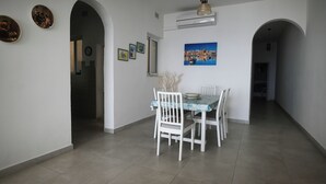 Un'area pranzo con un tavolo apparecchiato per quattro persone, sedie bianche e una tovaglia blu. Si notano tre quadri appesi al muro e un condizionatore sopra il tavolo.