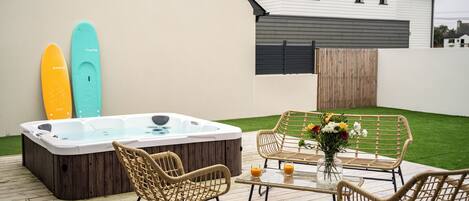 A modern outdoor patio with a hot tub, wicker furniture, and a table with drinks and flowers.