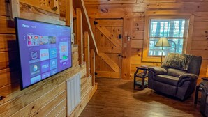 A wooden cabin interior with a mounted TV displaying Roku Home screen, a cozy seating area with a chair and table, and a large window with blinds. - Luxury cabin getaway on a 40 acre retreat (Bismarck)