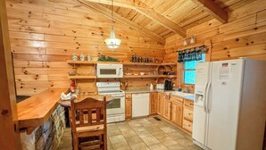 Une cuisine rustique, située dans une cabane en bois, avec un mur en pierre, des meubles en bois, un poêle blanc, un four à micro-ondes, un réfrigérateur et un luminaire suspendu.