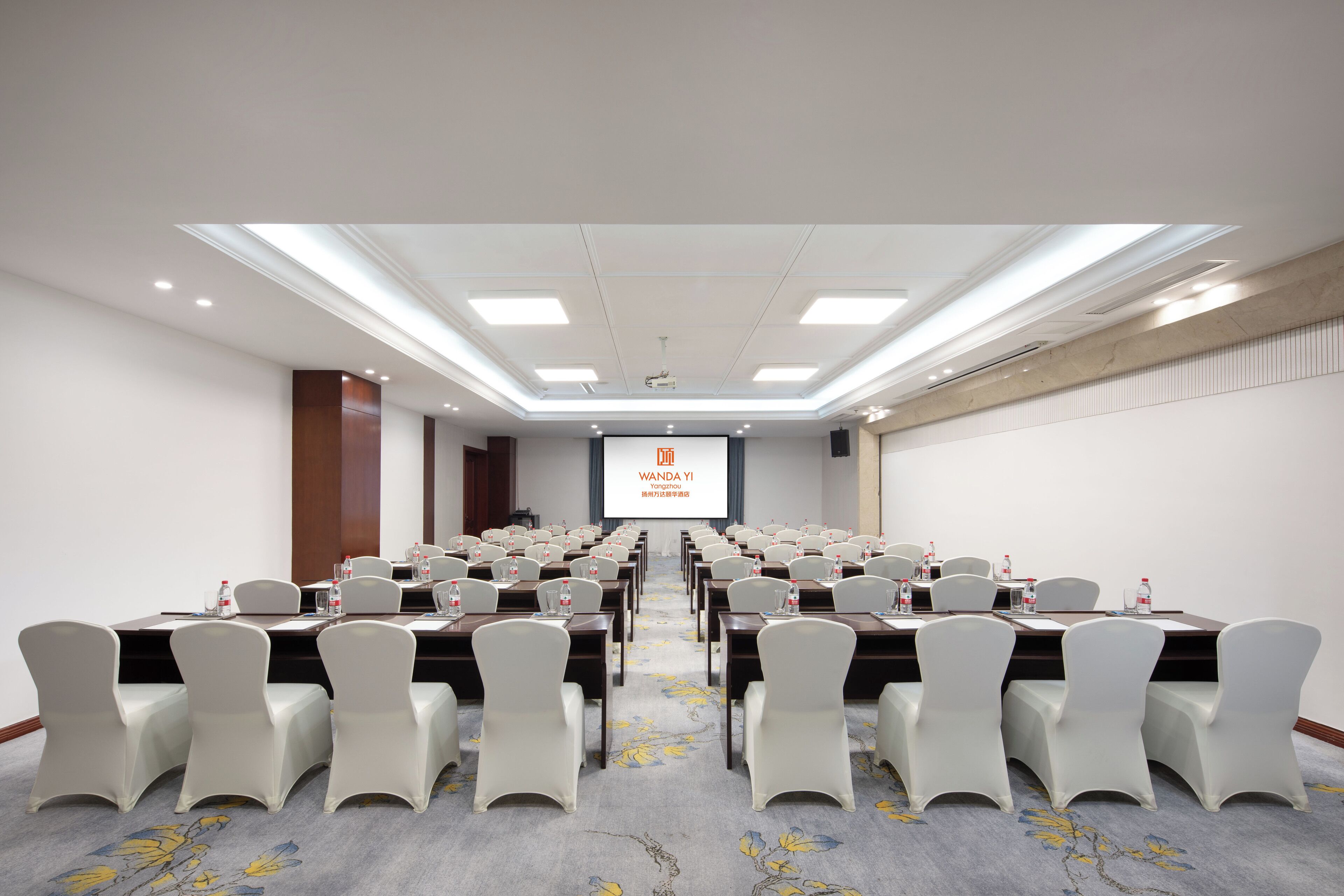 Un salón de conferencias con mesas y sillas dispuestas en filas, una pantalla de proyector y suelo alfombrado.