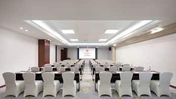 Un salón de conferencias con mesas y sillas dispuestas en filas, una pantalla de proyector y suelo alfombrado.