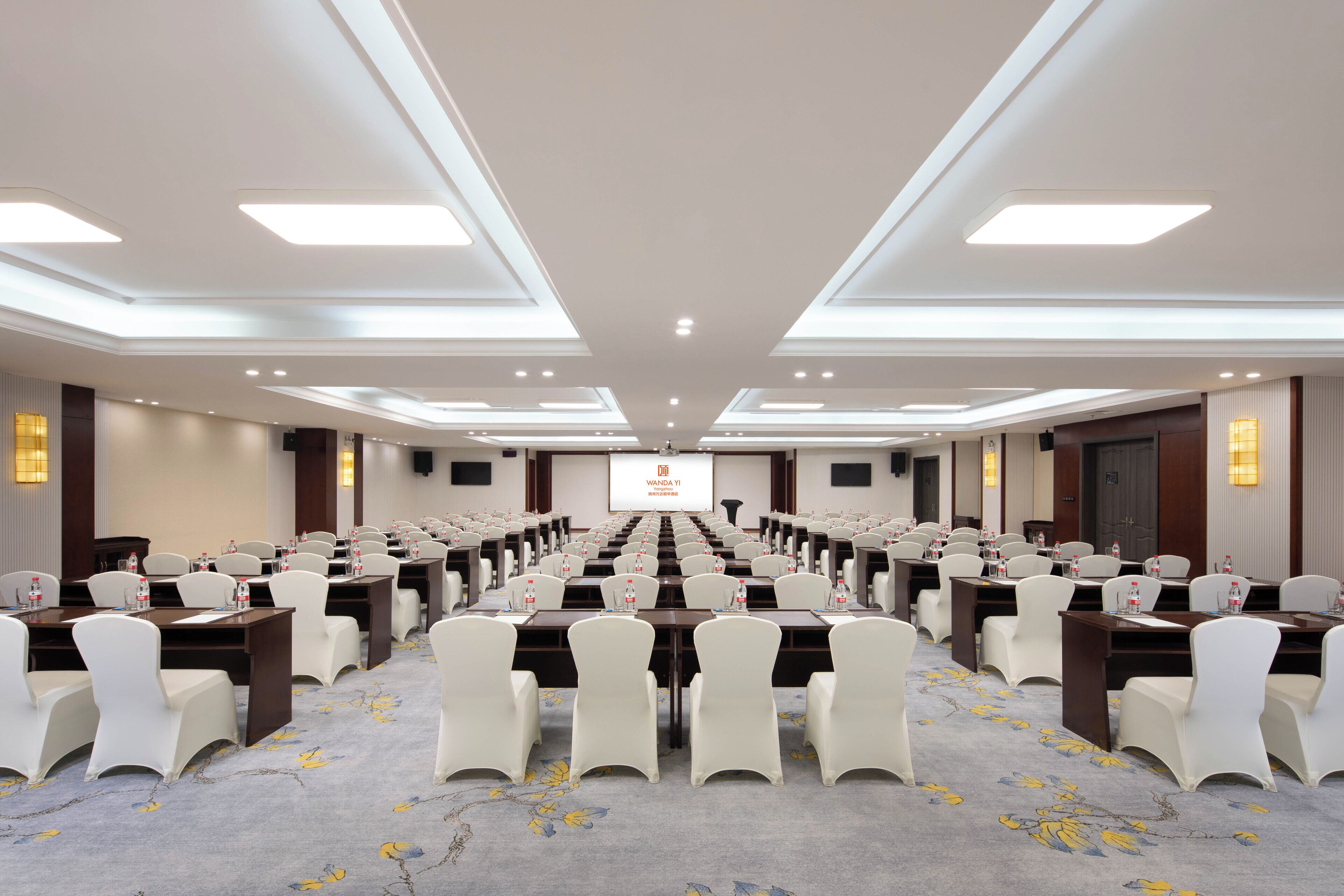 A conference room with rows of tables and chairs, a projector screen at the front, and a carpeted floor.