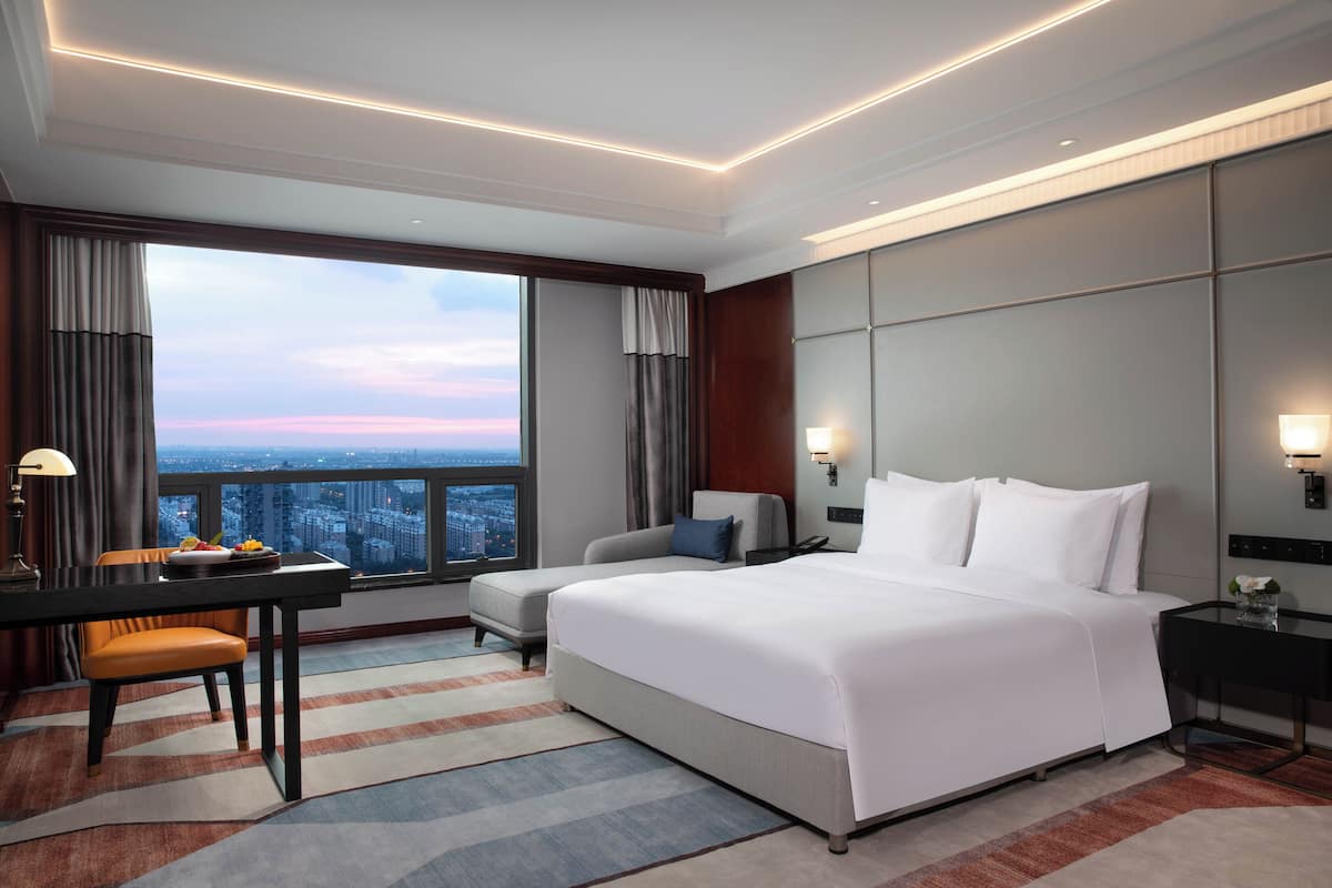 Een hotelkamer met een groot bed, een bureau met stoel, uitzicht op de stad door een groot raam en een nachtkastje met lamp.