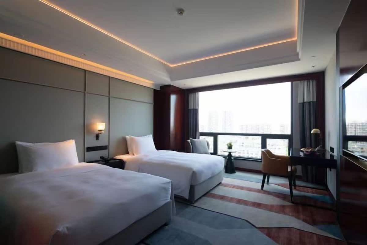 Een hotelkamer met twee bedden, een bureau, een stoel en een groot raam met uitzicht op de stad.