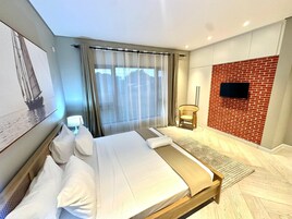 A modern hotel room with a large bed, a television mounted on a brick wall, and a window with sheer curtains.