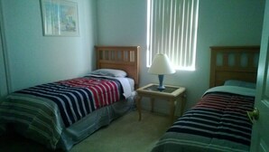 A small, simple room with two single beds, a nightstand with a lamp, and a window with blinds. - Calabay Parc - Spacious 4 bed to suit your family vacation (Davenport)