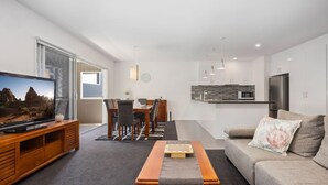 A modern living room with a flat-screen TV, a dining area with a wooden table and chairs, a kitchen with white cabinets and appliances, and a grey sofa with a floral pillow. - Stratton Summit 2, 5 bedroom townhouse in Jindabyne. (Jindabyne)