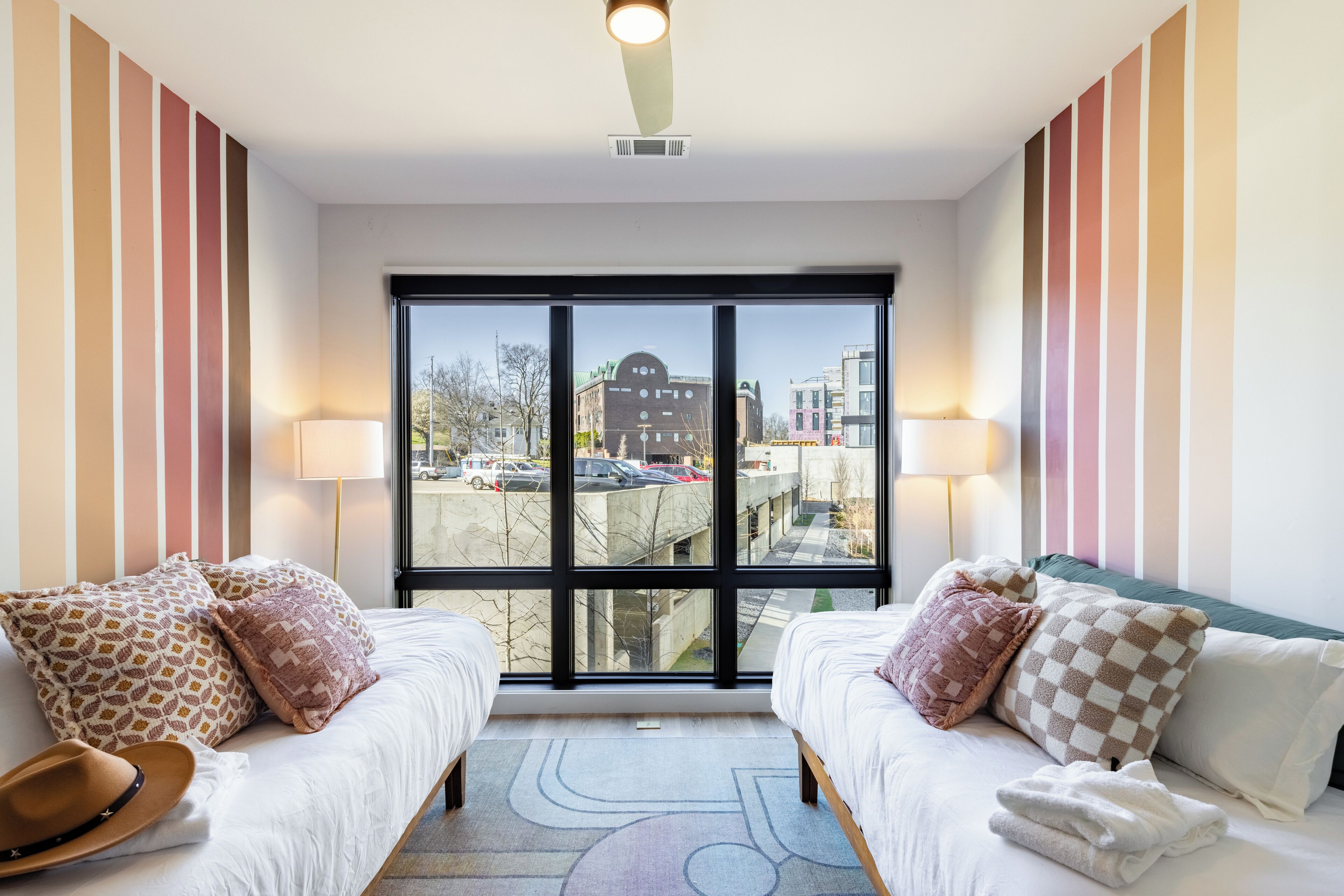 Ein Hotelzimmer mit zwei Betten, gestreiften Wänden, großen Fenstern und Blick auf die Stadt.