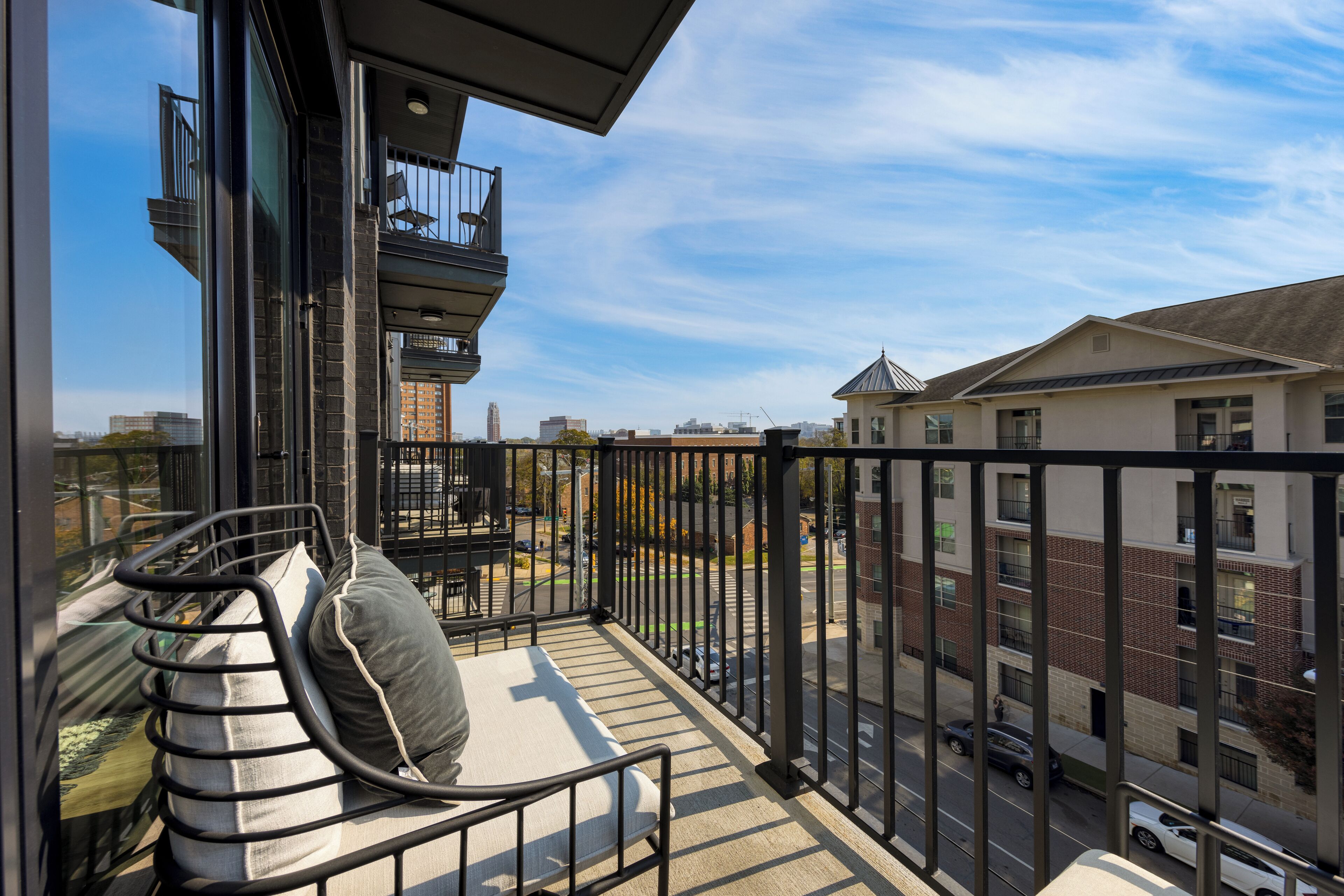 A balcony with a cushioned seating area, a view of the street, and a building with a brick facade in the background.