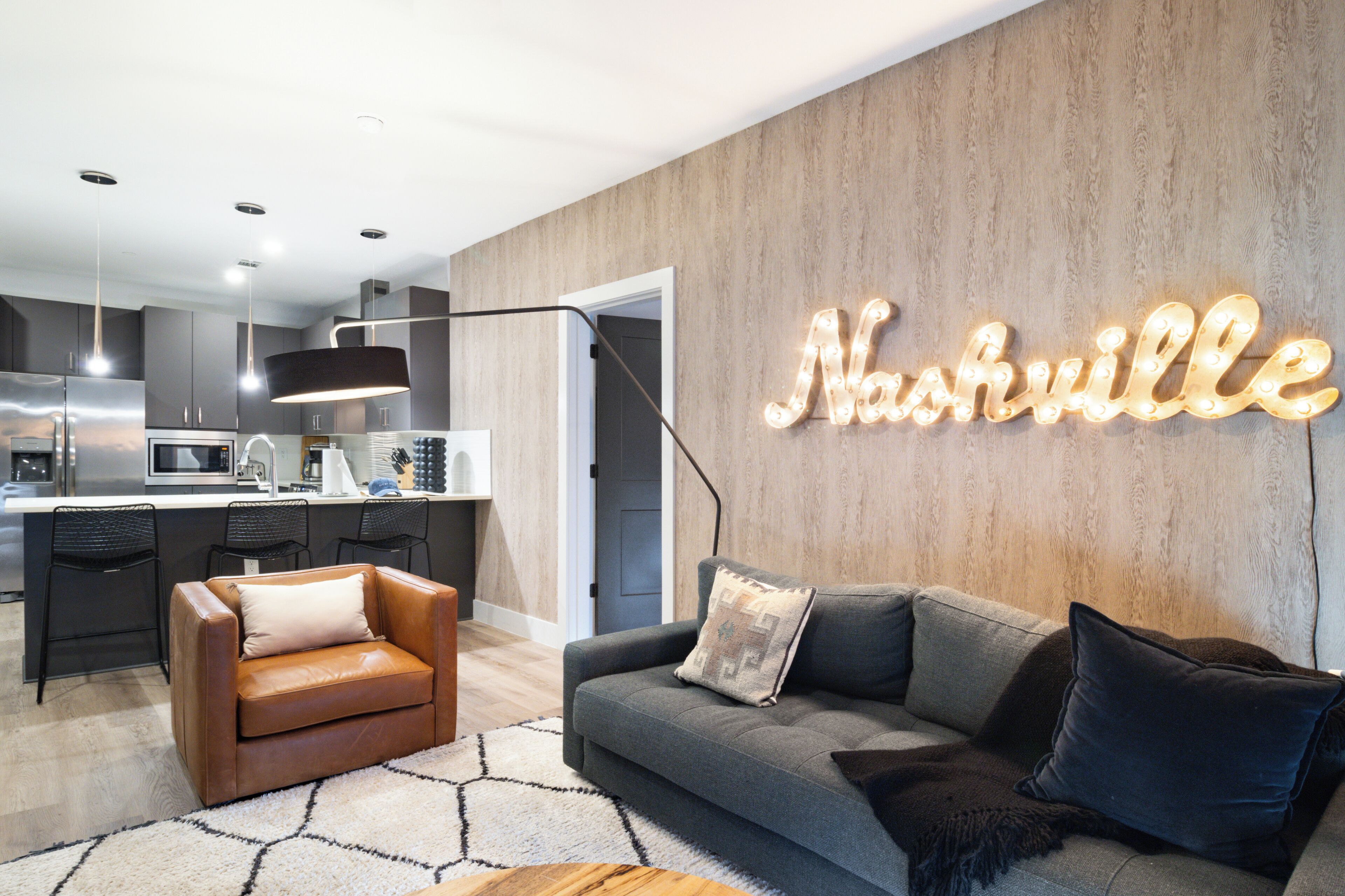Una sala de estar moderna con un sofá gris, un sillón de cuero marrón y una alfombra. Un letrero de neón en la pared dice "Nashville".