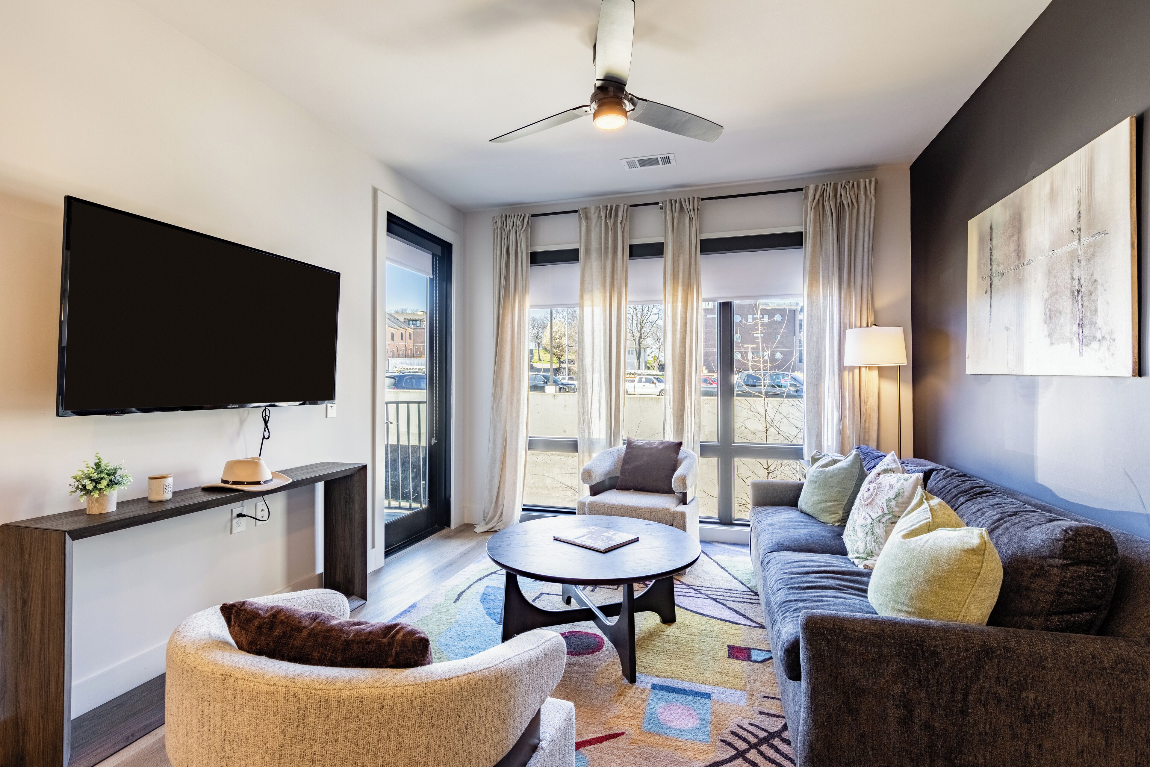 A modern living room with a flat-screen TV, a ceiling fan, a sofa, a coffee table, and a large window with curtains.