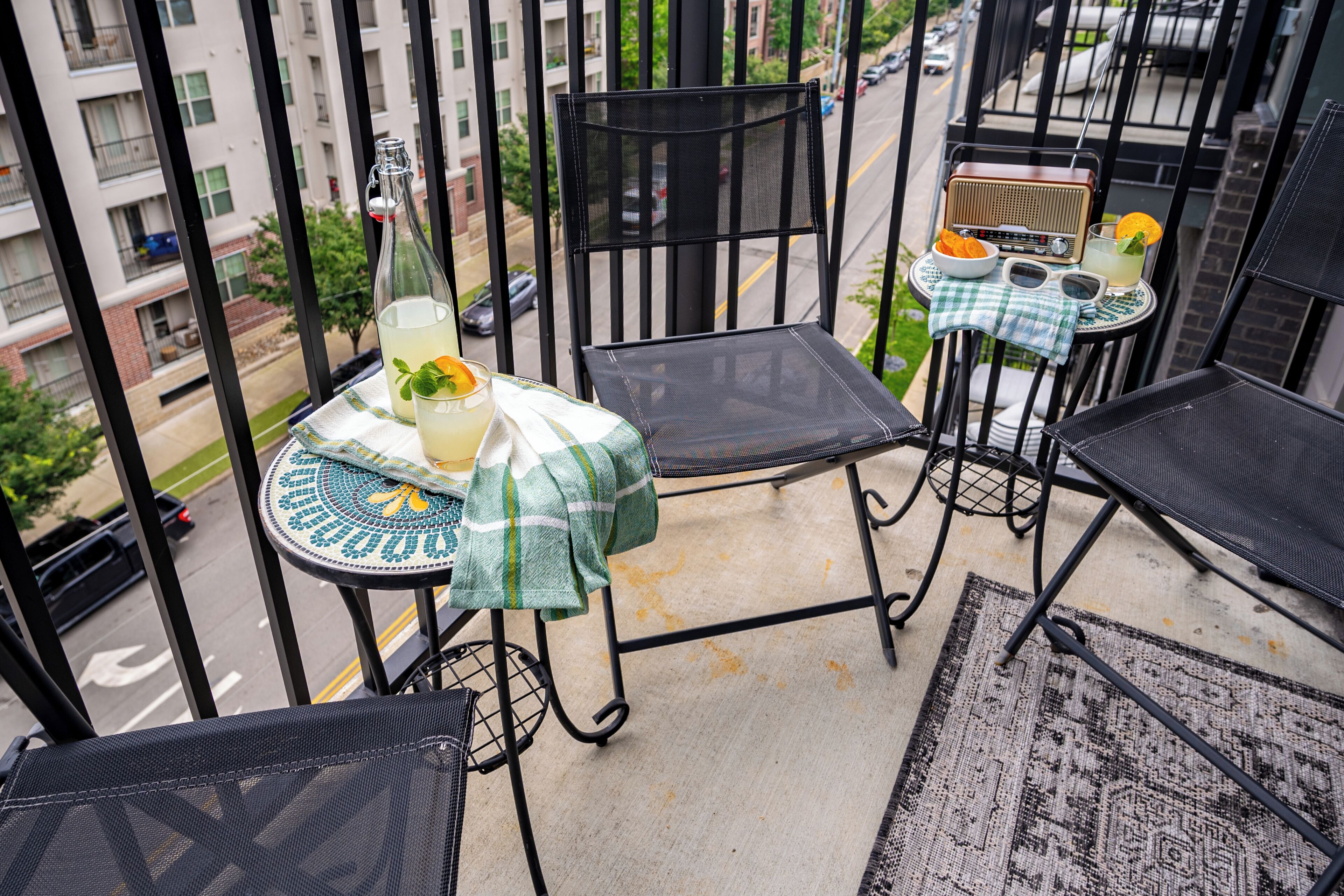 A balcony with two chairs, a small table with a checkered tablecloth, a bottle of water, a glass of lemonade, and a radio.