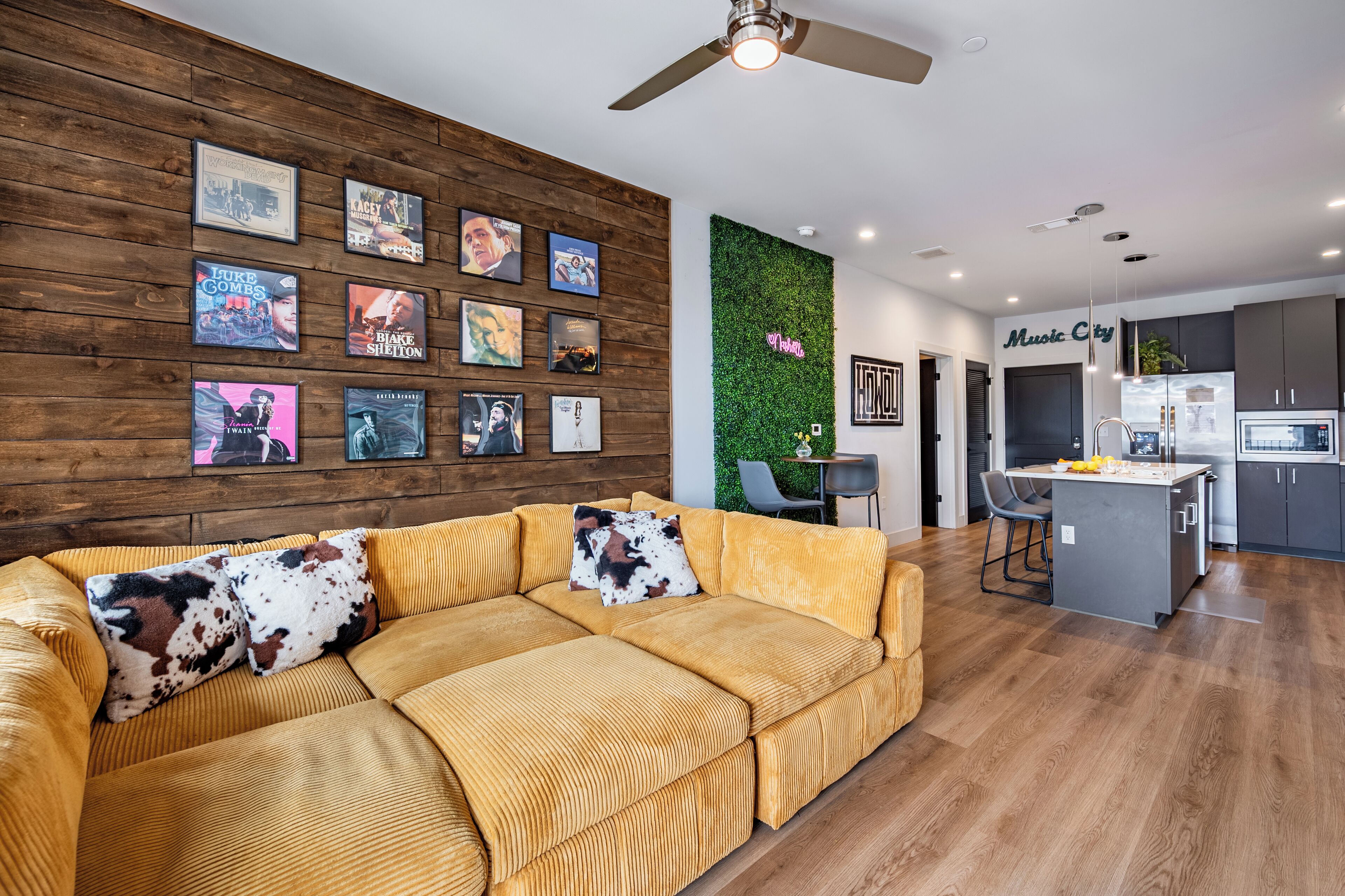 A modern living room with a large sofa, a wooden wall with framed movie posters, a green vertical garden, and a kitchen area with a bar.