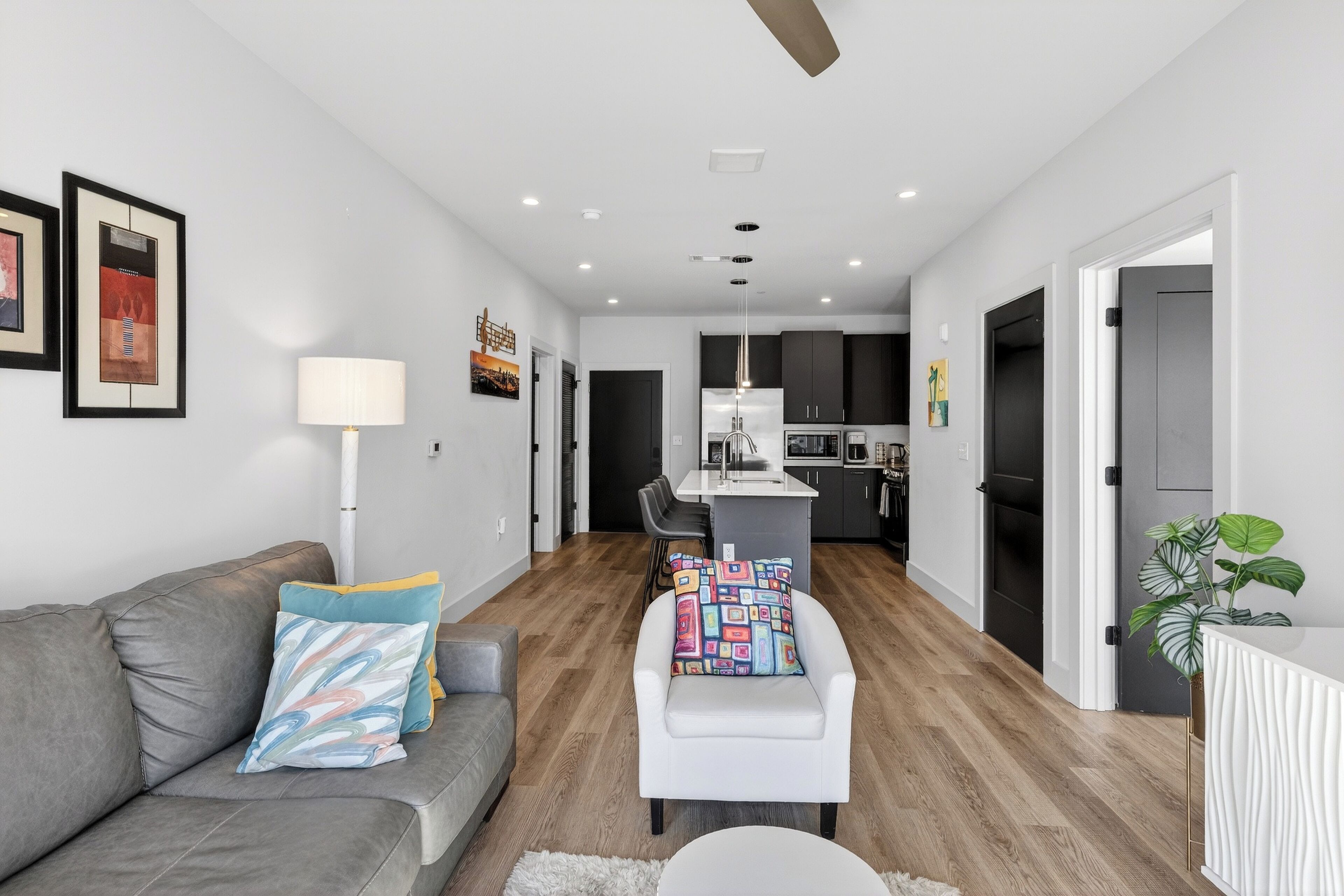 A modern living room with a grey sofa, a white armchair, and a small table. There is a kitchen in the background with dark cabinets and a white countertop.
