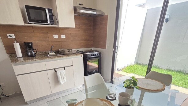 A modern kitchen with a microwave, coffee maker, and stove. There is a glass dining table with chairs and a view of the outdoors.