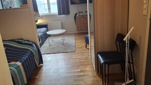 A compact living space with a bed, a small round table, a chair, a radiator, a TV, and a window with curtains.