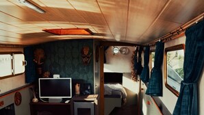 A narrow, well-lit interior with a wooden ceiling, a small TV on a table, a bed, and a window with curtains. - Unique nature experience on the water, Bed in boat Jisperveld (Spijkerboor)