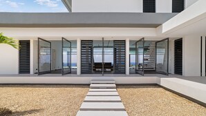 A modern house with a covered outdoor area, a gravel path leading to a seating area, and large glass doors. - Kas di Sol Infinito, NEW! Luxury Ocean View Villa w/ Huge Private Pool, 3 Bedrooms & Dive Facilities (Kralendijk)