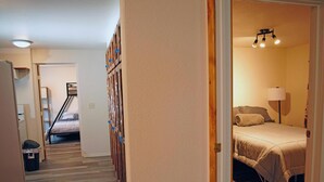 Un pasillo con suelo de madera conduce a un dormitorio con cama, mesita de noche y lámpara. Se ve una escalera al fondo.