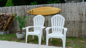 Two white plastic Adirondack chairs, a wooden surfboard, and a potted plant against a wooden fence.