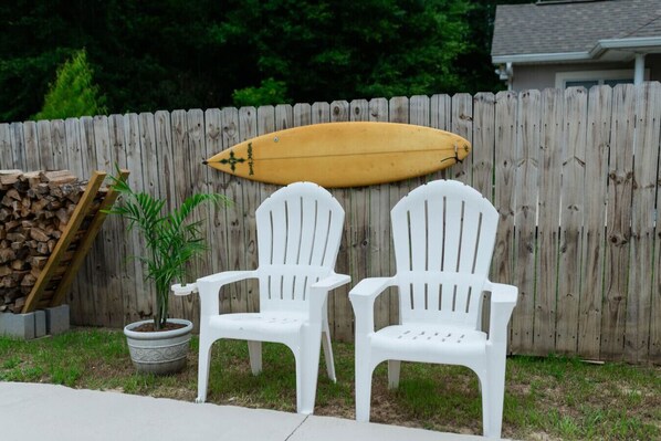 Dos sillas Adirondack blancas de plástico, una tabla de surf de madera y una planta en maceta frente a una valla de madera.