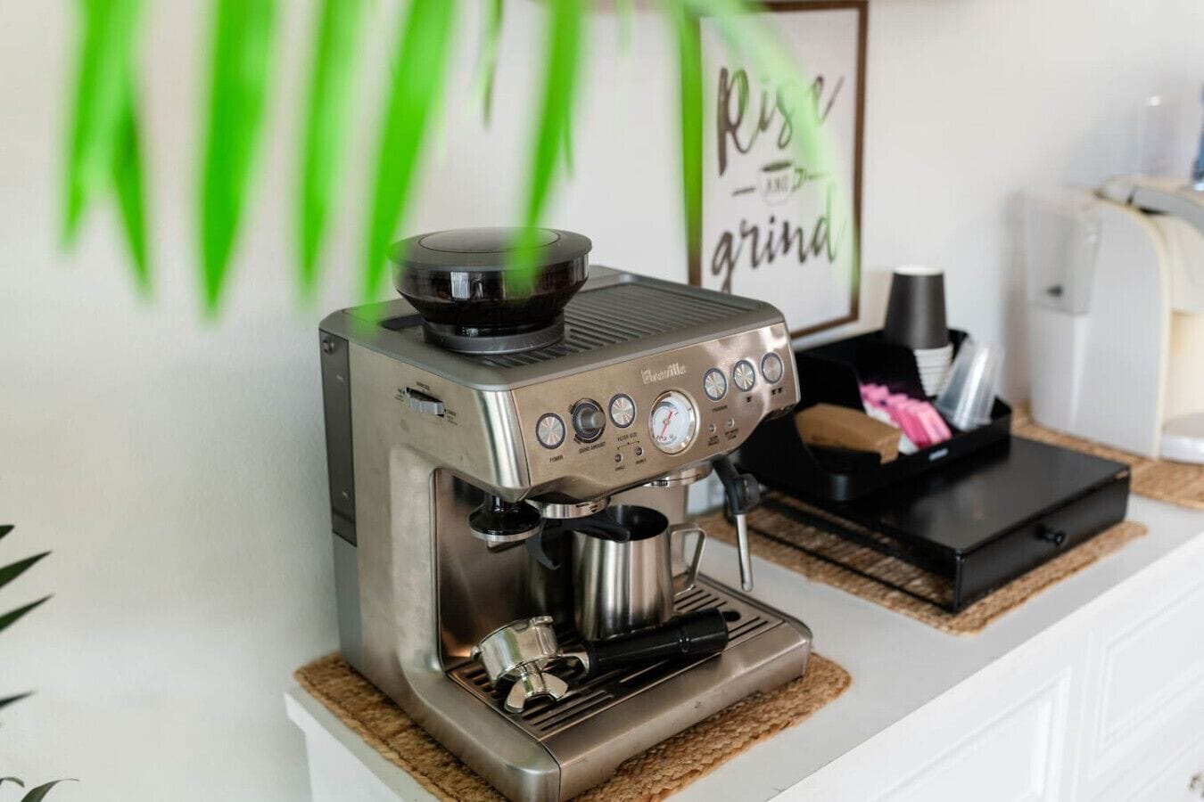 A coffee machine on a countertop with a coffee grinder and a framed sign in the background.