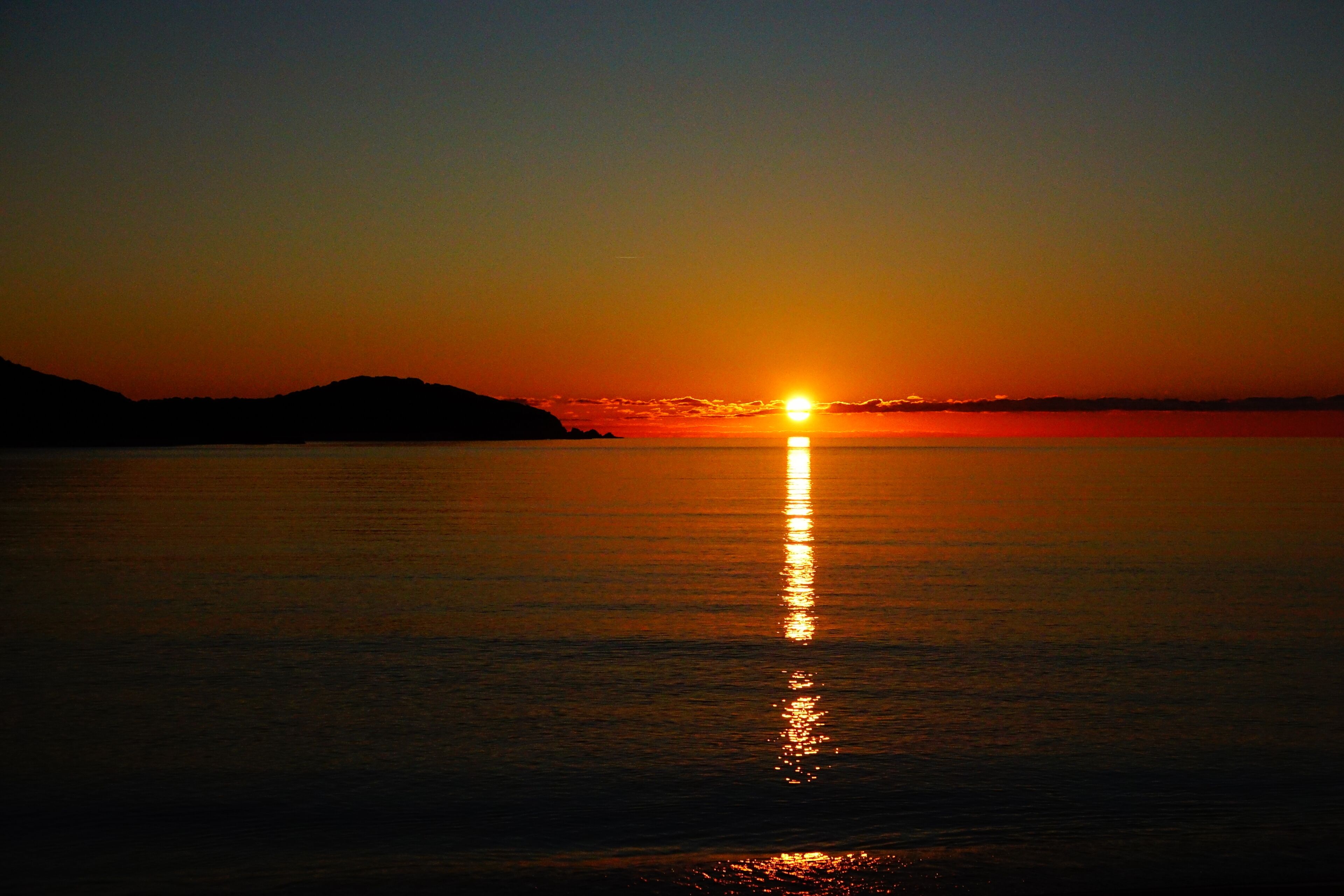 Ein Sonnenuntergang über ruhigem Wasser, im Hintergrund eine Bergkette als Silhouette.
