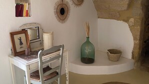 Een gezellige kamer met een wit bureau, een stoel, een klein plankje met boeken, decoratieve wanddecoratie, een groene vaas en een geweven vloerkleed.