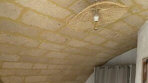 Een slaapkamer met een stenen plafond, een bed met witte lakens, een nachtkastje met een lamp en een geweven plafondlamp.