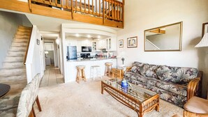 A spacious living area with a high ceiling, a kitchen with white cabinets, a wooden staircase, and a large mirror on the wall. -  2-bedroom apartment with WiFi, AC in Kihei (Kihei)