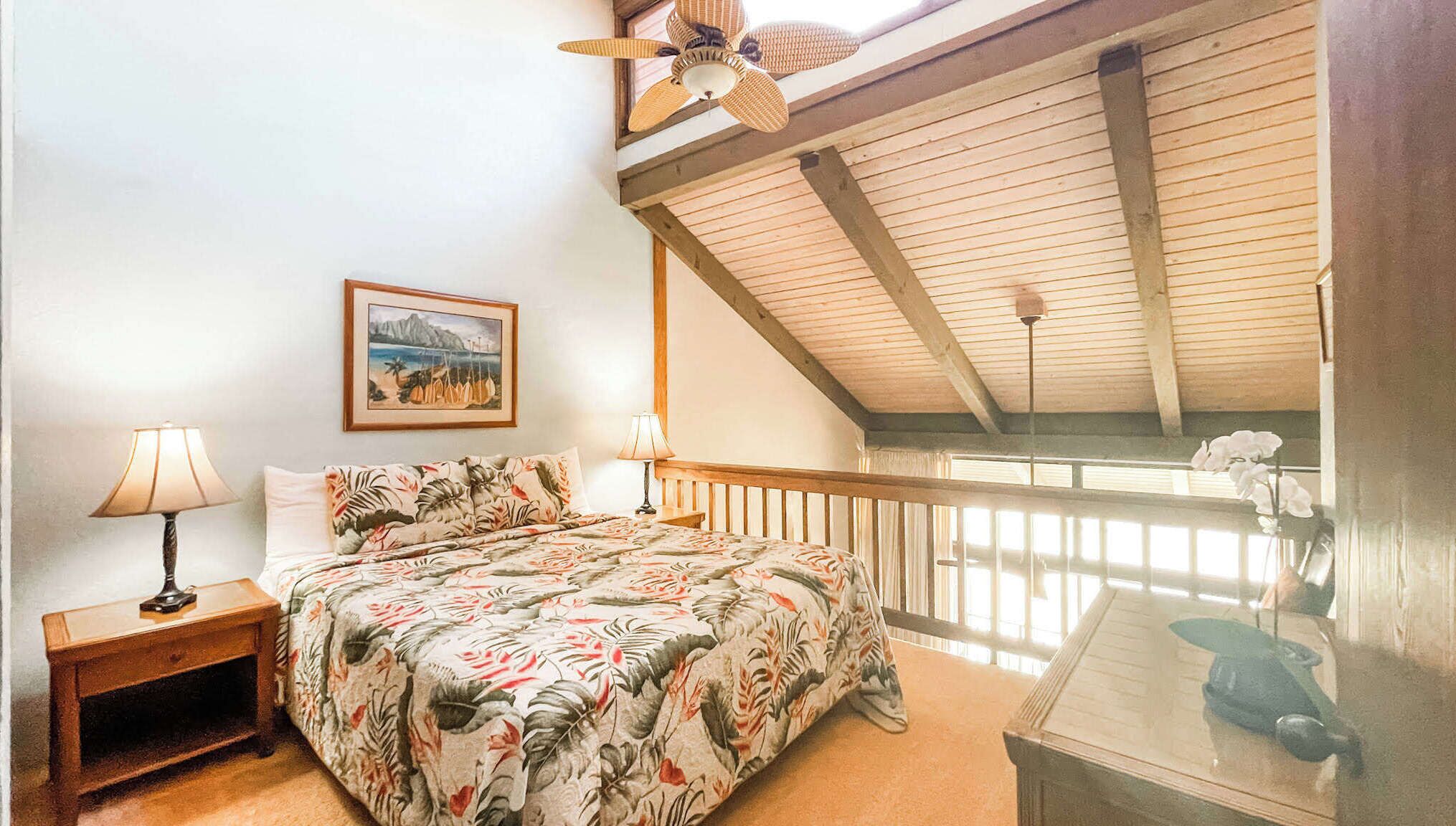 A bedroom with a bed, a nightstand, a lamp, a framed picture, a ceiling fan, and a balcony with a view.