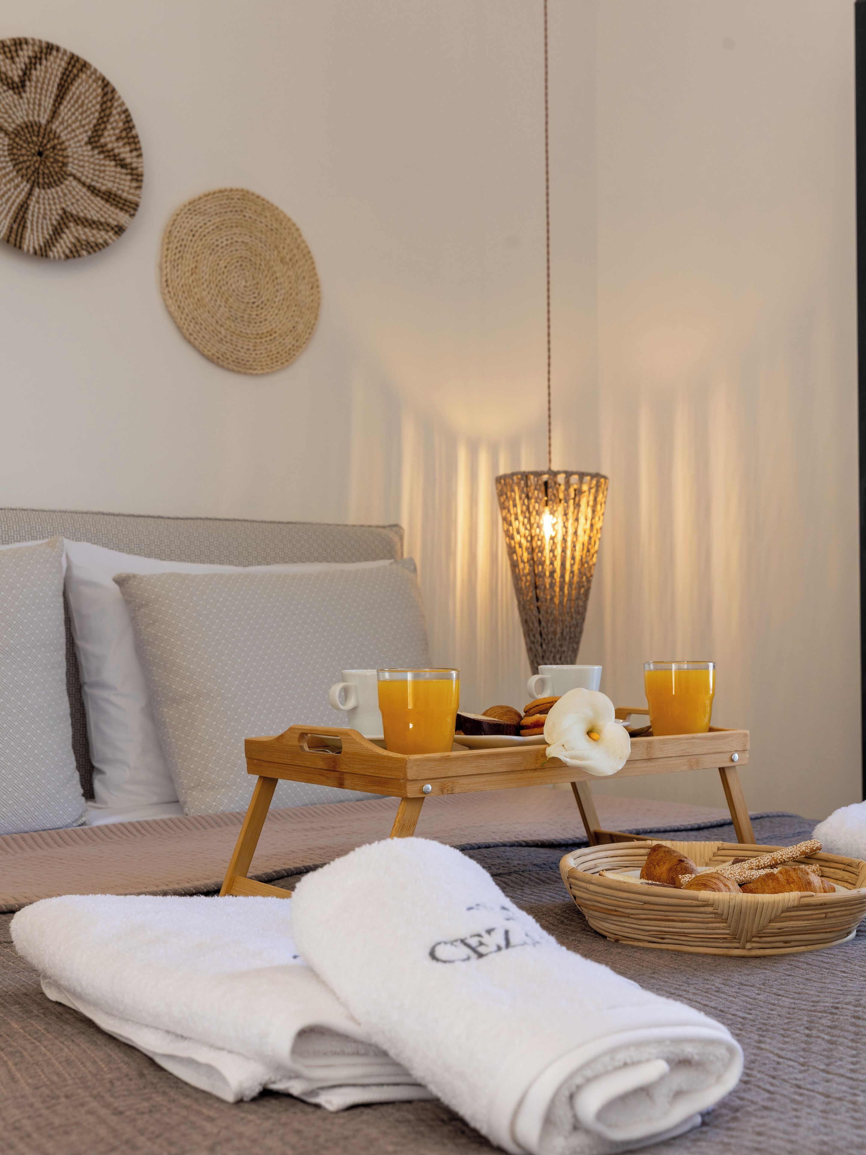 A neatly made bed with pillows, a tray with breakfast including croissants, orange juice, and a white flower, and a woven wall hanging.